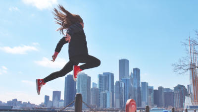 A woman wearing bright red vintage sneakers jumping with a city's skyline in the background.